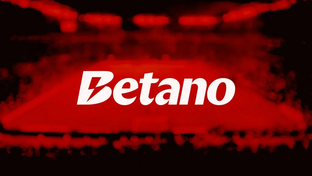 A imagem é um banner para a Betano. Diz Betano ao longo do centro. A imagem de fundo é de um estádio de futebol cheio de pessoas assistindo a um jogo. A imagem está borrada e é nas cores vermelha e preta.