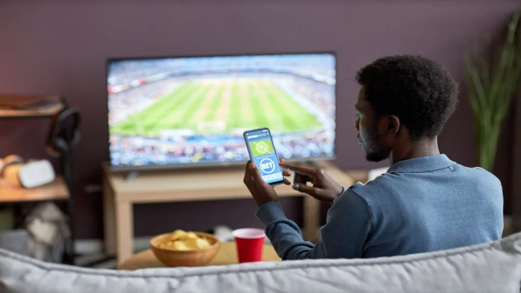 A imagem mostra um homem segurando um celular com o site da Betnacional, e ao fundo há uma tela com uma partida de futebol acontecendo.