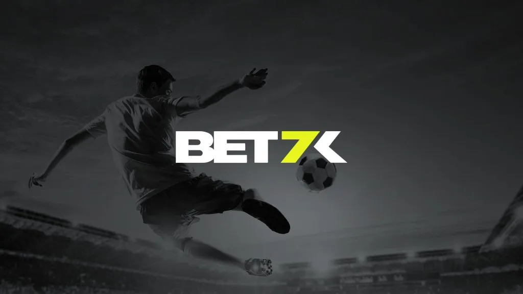 A imagem é um banner para a Bet7k. Diz Bet7k no centro. Ela está no branco e preto e mostra um jogador de futebol no ar, prestes a chutar uma bola em um estádio cheio de pessoas.