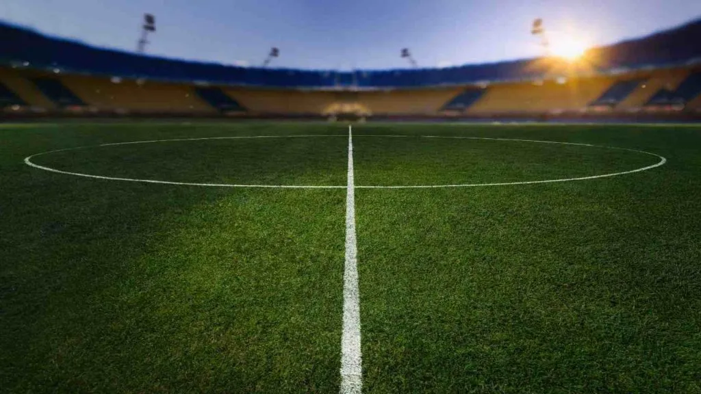 La foto muestra el centro de un campo de fútbol. Está tomada desde un costado siguiendo la línea que pasa por el centro. 