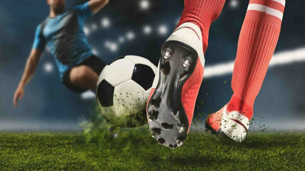 A imagem mostra um jogador de futebol prestes a chutar uma bola e outro no meio do salto, também tentando chutar a bola.