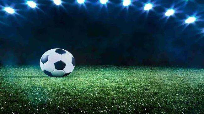 A imagem mostra uma bola de futebol no meio de um campo de futebol. O fundo é preto, mas a bola e o campo estão iluminados por um conjunto de refletores do estádio acima. 