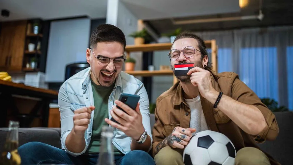 A imagem apresenta dois homens sentados no sofá apostando na Eurocopa na Betano.