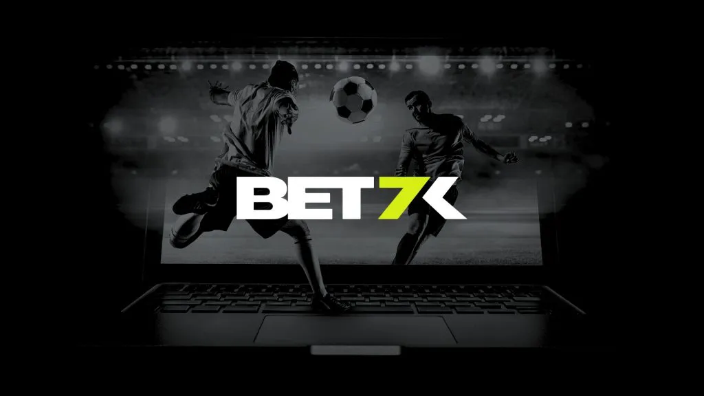 A imagem é um banner para a Bet7k. Diz Bet7k no centro. O fundo é preto e branco e mostra dois jogadores de futebol prestes a chutar uma bola que está no ar. Os jogadores estão em cima do teclado de um laptop.