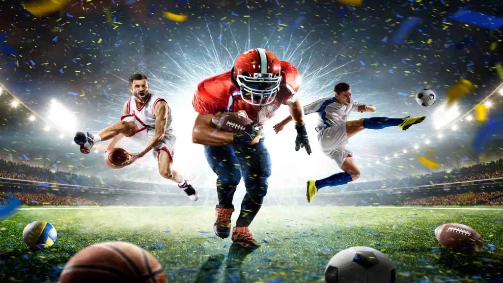A imagem mostra um jogador de futebol americano, um jogador de futebol e um jogador de basquete, todos no meio de um campo de futebol cheio de pessoas. Há confetes no ar e, na frente dos jogadores, há uma bola de vôlei, uma bola de basquete, uma bola de futebol e uma bola de futebol americano. Isso representa como a Betano oferece uma variedade de esportes para apostar.