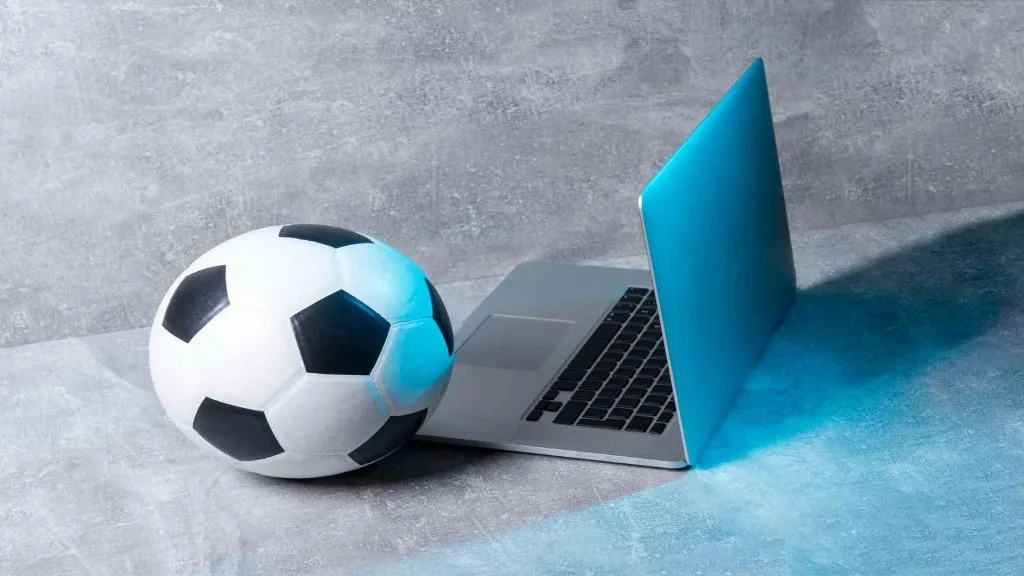 A imagem mostra uma bola de futebol ao lado de um laptop em um fundo cinza.