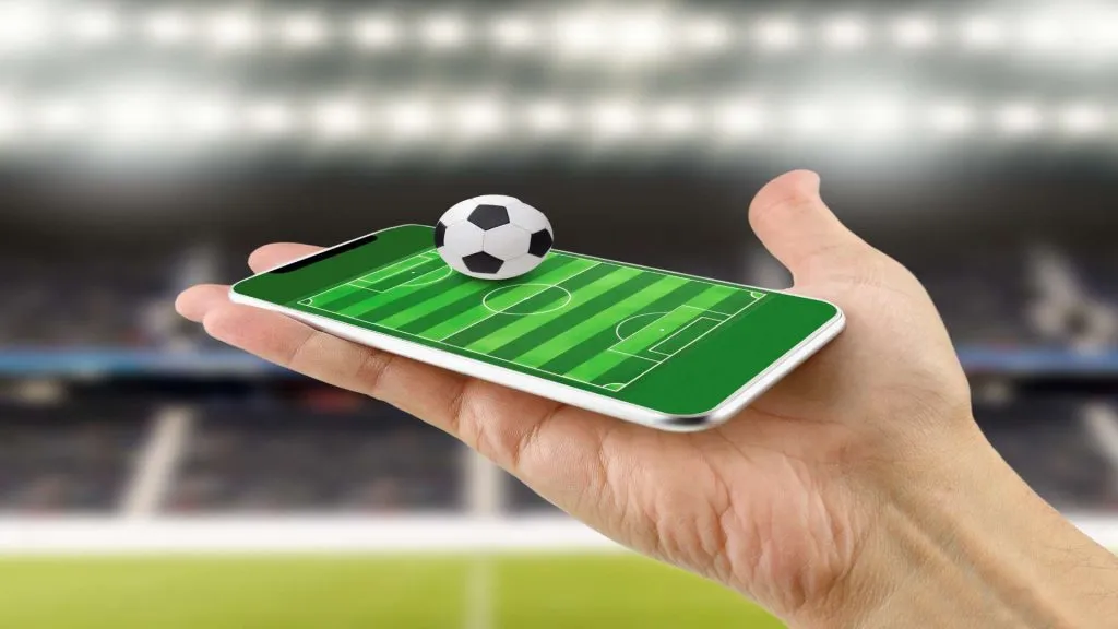 A imagem mostra uma mão segurando um celular, e a tela mostra um campo de futebol visto de cima. Em cima do celular, há uma bolinha de futebol preta e branca. 