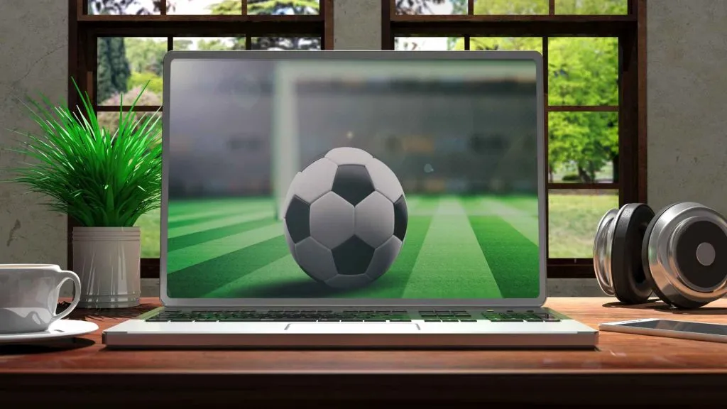 A imagem mostra um laptop em uma mesa junto a uma janela com uma xícara de café, uma planta e fones de ouvido ao lado. No laptop, há uma foto de uma bola de futebol no meio de um campo de futebol prestes a ser chutada para o gol.