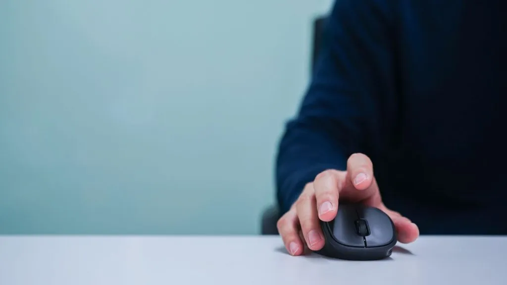A imagem mostra um homem sentado em uma mesa usando um mouse, em representação de como acessar o login da Luva Bet.