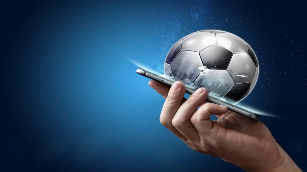 A imagem mostra uma mão segurando um celular com uma bola de futebol preta e branca saindo dele.