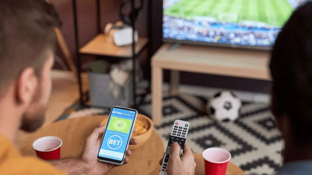 A imagem mostra dois homens assistindo a um jogo de futebol, e um deles está com um celular com a Globo bet na tela.
