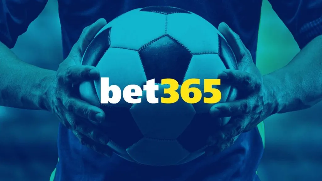 A imagem é um banner para a bet365. Diz bet365 no centro e o fundo mostra um jogador de futebol segurando uma bola de futebol preta e branca nas mãos. A imagem de fundo também tem um filtro azul.