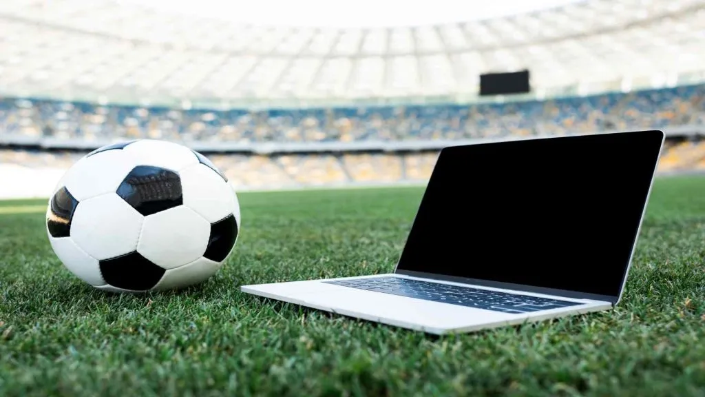 A imagem mostra uma bola de futebol preta e branca e um laptop no meio de um campo de futebol em um estádio.
