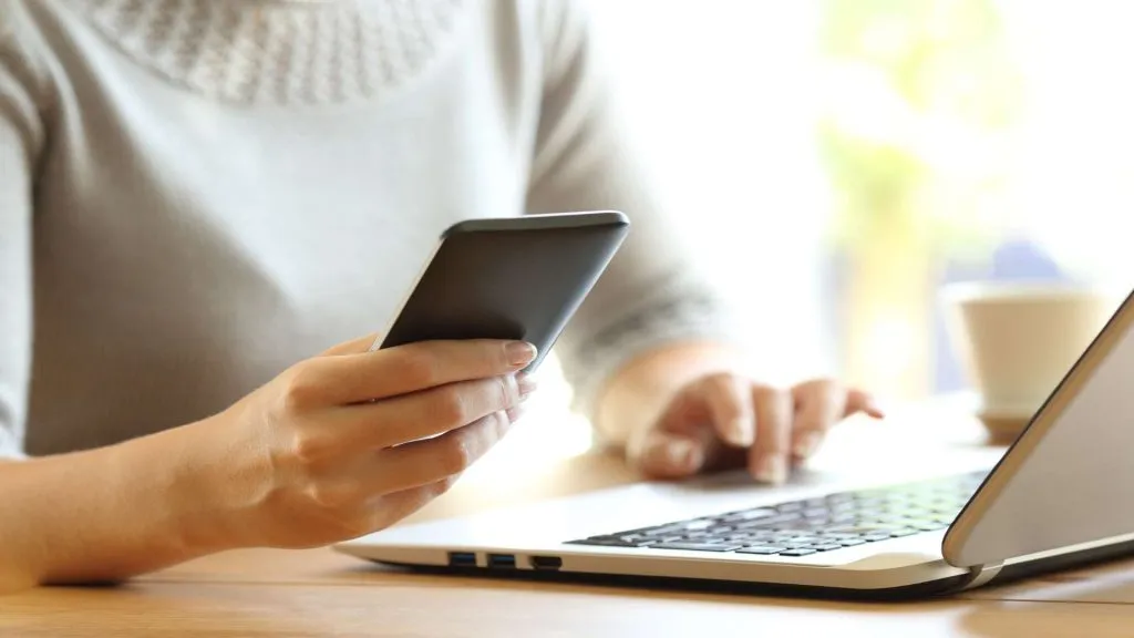 A foto é de uma mulher usando o celular e o laptop, representando que é possível acessar a Aposta Ganha Brasil por diferentes dispositivos.