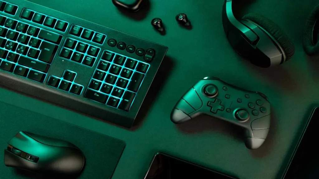 A imagem mostra um conjunto de fones de ouvido e um headset, um controle de videogame, um teclado e um mouse, todos em cima de uma mesa. A imagem é principalmente nas cores verde e preto.