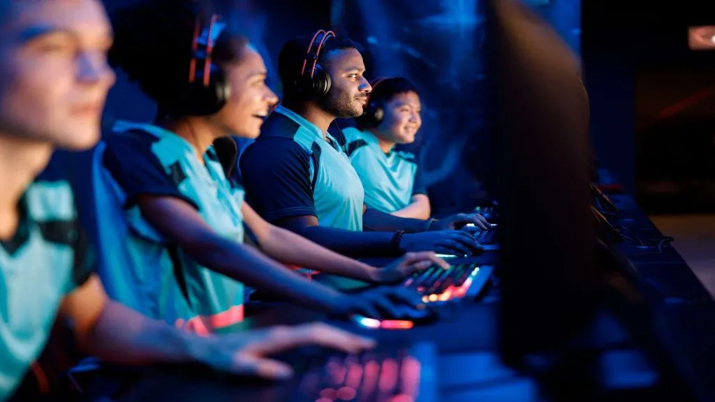 A imagem mostra quatro pessoas jogando eSports em computadores. Todos estão vestindo camisetas azuis idênticas, o que indica que todos podem ser do mesmo time.