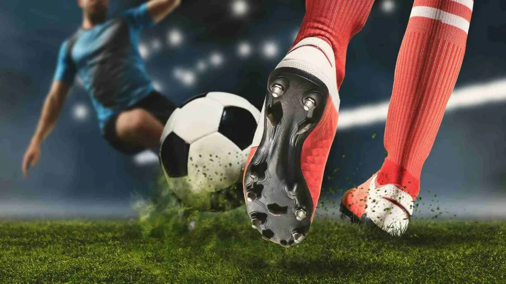 A imagem mostra um jogador de futebol prestes a chutar uma bola e outro no meio do salto, também tentando chutar a bola.