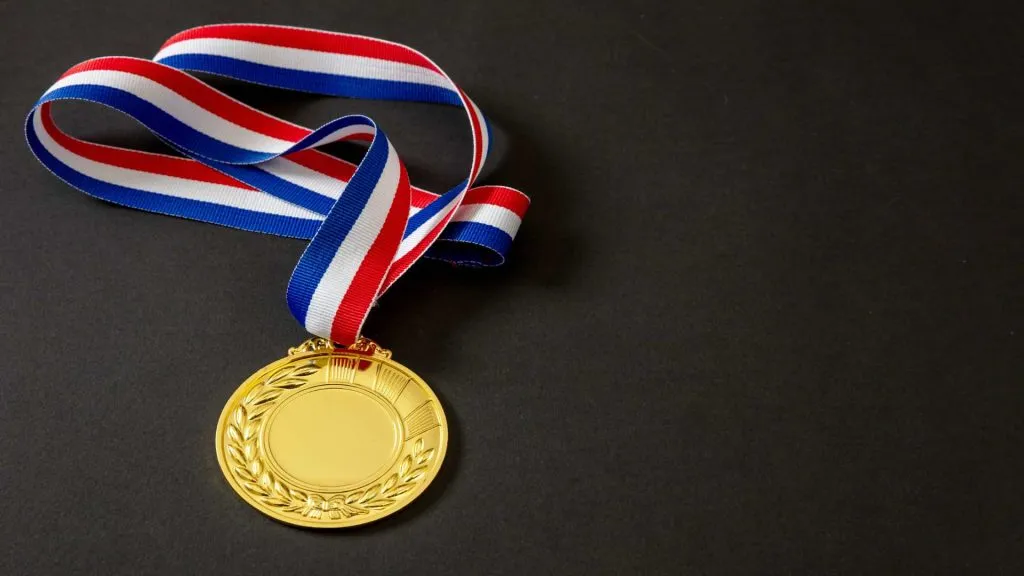 A imagem mostra uma fita listrada segurando uma medalha de ouro sobre uma mesa. O fundo é preto.