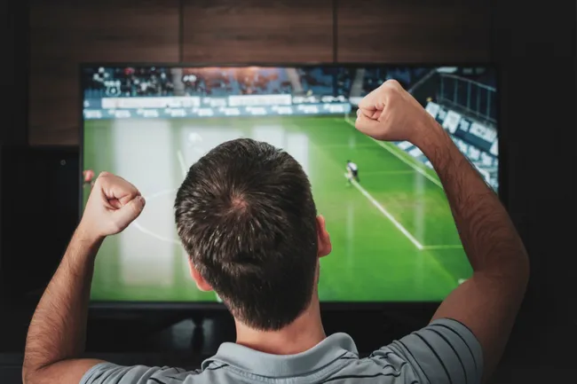 A imagem mostra um homem assistindo a um jogo de futebol pela TV e torcendo, representando que a Superbet Brasil tem os principais esportes e campeonatos do mundo para apostar ao vivo. 