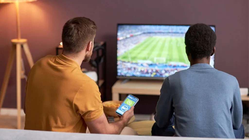 A imagem apresenta dois homens sentados no sofá e olhando um game de futebol fazendo apostas ao vivo na Sportingbet. 