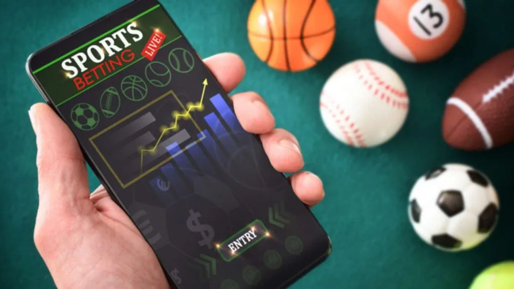 A imagem mostra uma mão segurando um celular com um site de apostas na tela. Ao fundo, há bolas para diferentes esportes, como basquete, futebol, tênis, beisebol, futebol americano e sinuca. Isto representa que Stake conta com mercados para apostar ao vivo em esportes. 