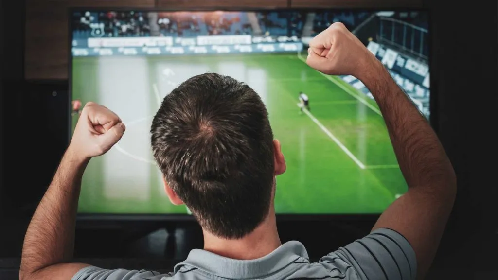 A imagem mostra um homem assistindo a uma partida de futebol na TV e torcendo.