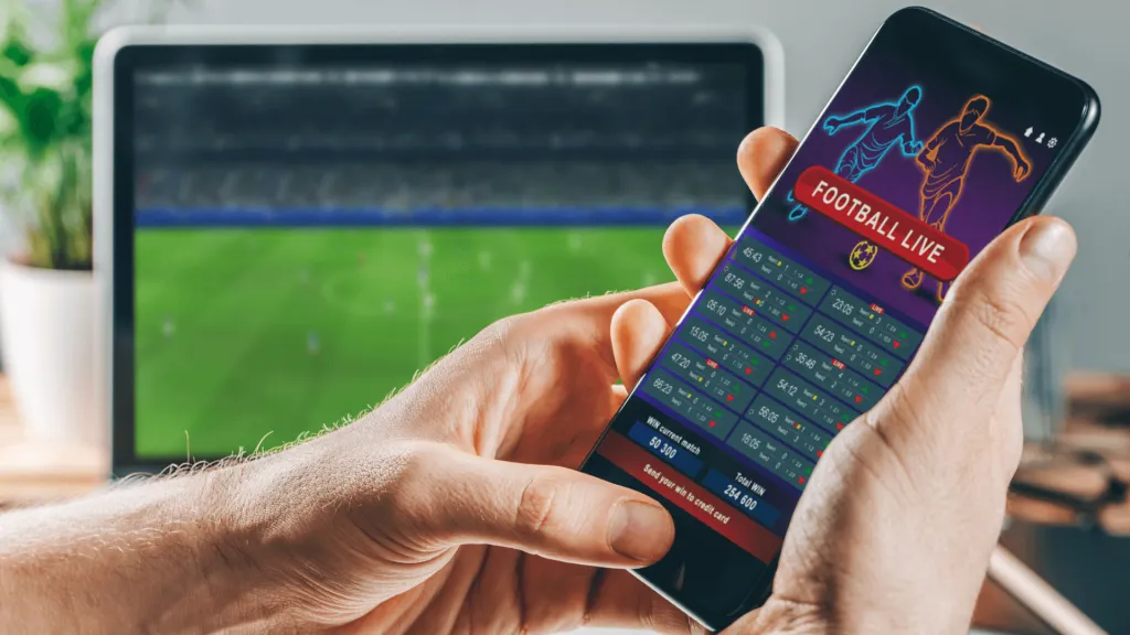 A imagem mostra um homem segurando um celular com uma casa de apostas aberta, e ao fundo há uma tela com uma partida de futebol acontecendo.