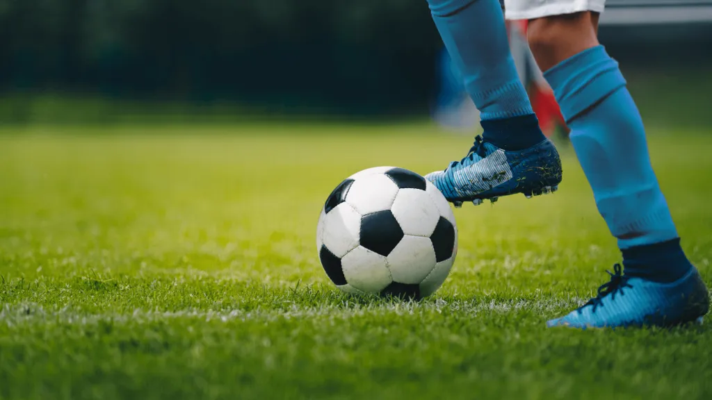 Esta imagem mostra um jogador com uma bola de futebol no campo, a foto foi tirada dos joelhos para baixo, destacando a bola como ponto focal.