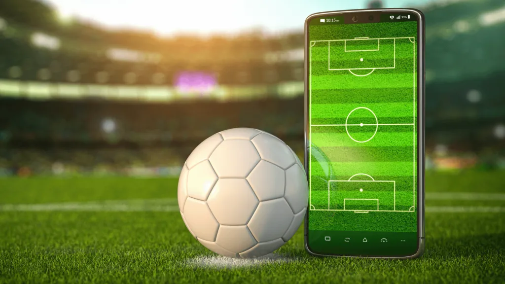 A imagem mostra uma bola de futebol totalmente branca no meio de um campo de futebol ao lado de um celular, que também mostra um campo de futebol de cima, em representação do app da Bet dá Sorte.