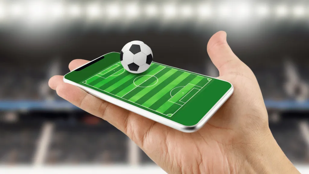 A imagem mostra uma mão segurando um celular, e a tela mostra um campo de futebol visto de cima. Em cima do celular, há uma bolinha de futebol preta e branca.